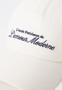 Crème baseballpet gemaakt van gestructureerde stof, met geborduurde marineblauwe tekst "L'école Parisienne de L'amour Moderne" op de voorkant.