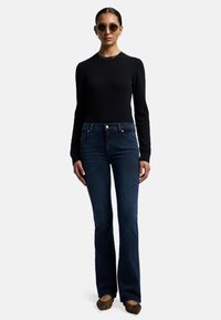 Svart långärmad tröja, mörkblå utsvängda jeans, runda solglasögon och mönstrade flats. Mörka, släta texturer med avslappnad passform. Enfärgad bakgrund.