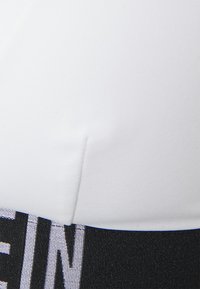Hvit sports-bh med svart elastisk bånd som har hvit skrift. Glatt materiale, tettsittende design, og kontrasterende farge detaljer.