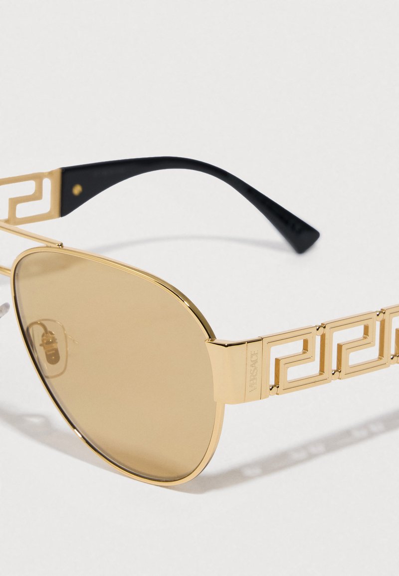 Gafas de sol aviador doradas con lentes marrones, varillas negras y un distintivo patrón de llave griega en los laterales. Materiales de metal y plástico utilizados.