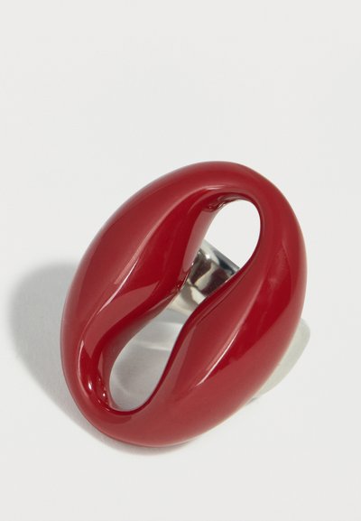 Bague ovale rouge brillante avec un centre creux et un design courbé lisse sur une bande argentée, photographiée sur une surface blanche.