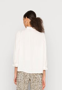 Blusa blanca con cuello fruncido, mangas abullonadas y corte recto. Combinada con pantalones de estampado de leopardo. Textura suave.
