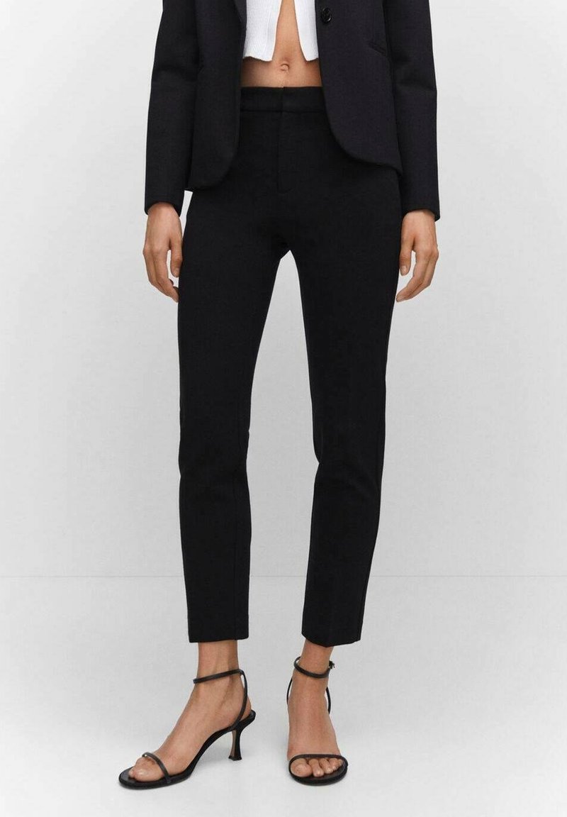 Pantalon noir sur mesure à coupe slim, longueur raccourcie et texture lisse, associé à des sandales à talons noires avec brides.