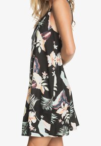 Femme portant une robe noire sans manches avec un grand imprimé floral pastel et des feuilles, debout de profil les mains derrière le dos.