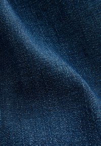 Tessuto di denim blu scuro con una superficie leggermente testurizzata, caratterizzato da un'intessitura uniforme e una lieve lucentezza. Nessun motivo o dettaglio distintivo visibile.