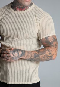 Kremowy t-shirt z dzianiny o ribbowanej fakturze, krótki rękaw i okrągły dekolt. Posiada dopasowany krój oraz subtelne, pionowe linie.