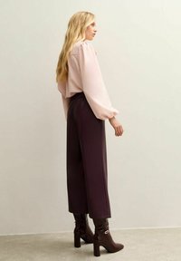 Light pink, puff-sleeve blouse paired with dark purple, wide-leg trousers. Model wears brown, heeled boots with a decorative strap.