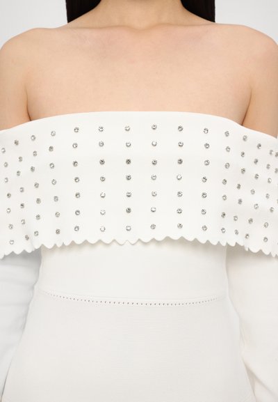Witte off-the-shoulder top met een gegolfde zoom, versierd met zilveren studs, zachte textuur en getailleerde silhoet. Gegolfde details aan de onderkant.
