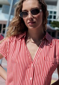 Vörös-fehér csíkos rövid ujjú ing zsebbel, könnyű anyagból, gombos záródással. Ovális napszemüveggel viselve.