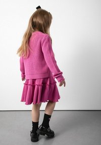 Różowy sweter z dzianiny o prążkowanej fakturze, zestawiony z różową sukienką plisowaną z falbaniastymi warstwami. Czarne skarpetki sięgające kostek i błyszczące czarne buty.