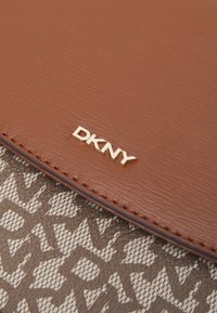 Brązowa skórzana torebka z złotym logo "DKNY". Tło stanowi jasna tkanina z powtarzającym się ciemnym wzorem tekstowym.