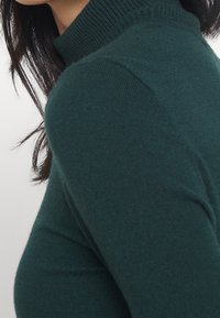 Pull à col roulé vert foncé, texture côtelée au niveau du col, coupe ajustée mettant en valeur les épaules et l'encolure, tissu doux, sans ornements.