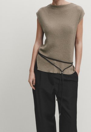 Ærmeløs beige striktop kombineret med sorte bukser med vide ben, med et tyndt sort omslagsslips bundet om taljen.