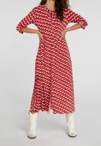 Robe rouge à manches longues avec un motif géométrique dans des nuances de rouge et blanc. Comprend un col, une fermeture à boutons, et une jupe plissée.