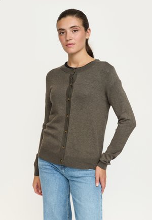 Femme portant un cardigan marron boutonné à manches longues et un jean bleu clair, debout avec une main sur la hanche devant un fond uni.