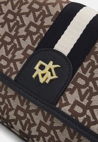 Brązowa i beżowa torba z tkaniny w wzory, z czarnym paskiem, teksturowanym złotym logo oraz gładką fakturą ze kontrastującymi akcentami.