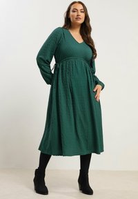 Långärmad, V-ringad grön klänning med texturerad tyg, samlad midja och knytning. Matchad med svarta ankelskor.