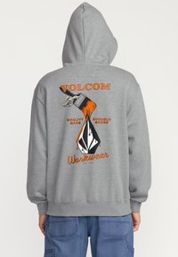 Grijze hoodie met een oranje en zwart grafisch ontwerp op de achterkant, met een hand die een verfkwast vasthoudt en tekst. Zachte stof.