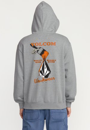 Grijze hoodie met een oranje en zwart grafisch ontwerp op de achterkant, met een hand die een verfkwast vasthoudt en tekst. Zachte stof.