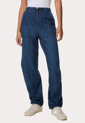 Žena oblečená v tmavě modrých volných džínových kalhotách s předními našitými kapsami, stahovací šňůrkou v pase a bílými teniskami.