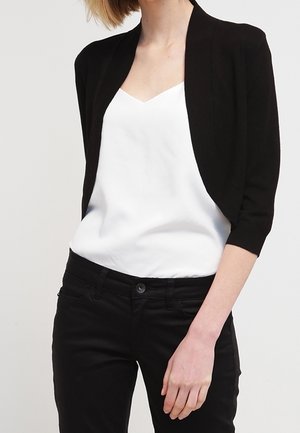 Kvinde iført en sort kort cardigan over en hvid V-hals camisole og sorte bukser mod en ensfarvet baggrund.