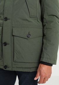 Veste matelassée verte avec une finition texturée, dotée de boutons noirs, de deux grandes poches avant et d'une fermeture éclair.