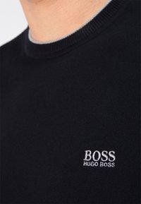 Pull noir en maille avec un col rond, présentant un col côtelé gris. Logo brodé "BOSS HUGO BOSS" en argent sur la poitrine.