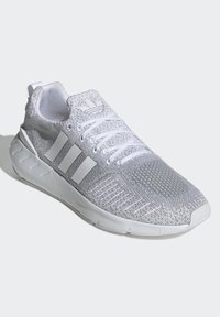 Fehér sportcipő, texturált hálós felsőrésszel, három fehér csíkkal és formázott gumitalppal, amelyen hangsúlyozott barázdák találhatók.