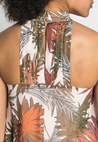 Top sin mangas floral con un cuello alto, que presenta una espalda plisada y transparente. Los colores incluyen naranja, verde y marrón sobre un fondo claro.