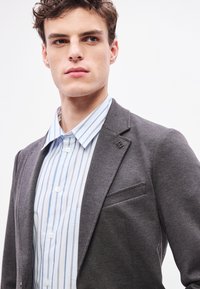 Γκρι σακάκι με notch πέτο και τσέπη, σε συνδυασμό με γαλάζιο ριγέ πουκάμισ�ο με γιακά με κουμπιά και λευκά κουμπιά.