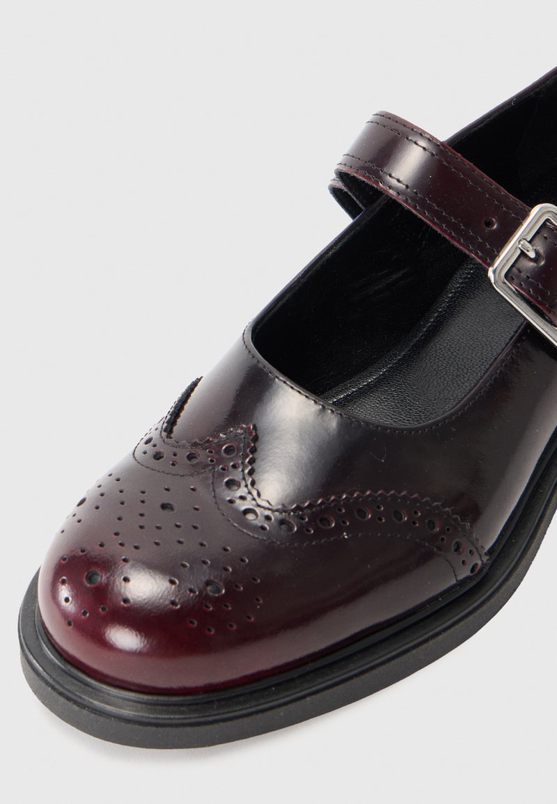 Chaussure Mary Jane en cuir bourgogne foncé avec des détails de brogue perforés, une semelle en caoutchouc noire et une seule sangle ajustable avec une boucle en métal.