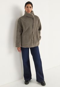 Sak fuzzy berwarna taupe dengan kerah tinggi, penutupan depan, dan kantong samping dipadukan dengan celana denim gelap berpotongan lebar.