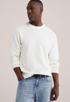 Homme portant un pull blanc à manches longues et un jean bleu clair avec une ceinture tressée marron, regardant sur le côté, mains dans les poches.