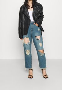 Giacca da moto in pelle nera indossata sopra una canotta bianca, abbinata a jeans blu strappati con grandi lacerazioni e orli sfrangiati. Sandali neri con cinturini.