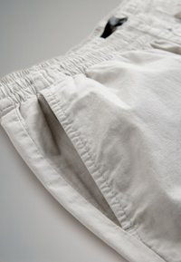 Gros plan sur un pantalon en coton beige avec taille élastique et détail de poche latérale sur un fond neutre.