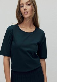 Donkergroen shirt met korte mouwen en een ronde hals, gemaakt van een gladde stof; een zwarte ribgebreide rok is zichtbaar ter hoogte van de taille.