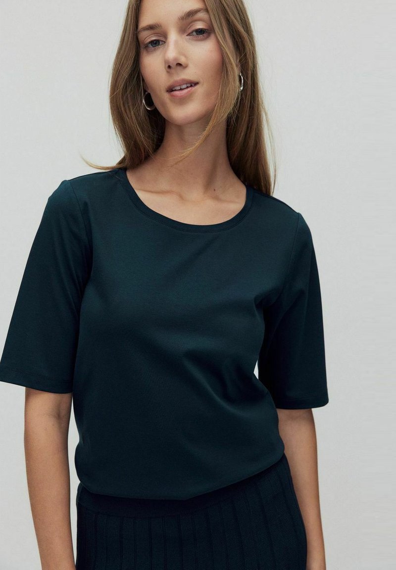 Donkergroen shirt met korte mouwen en een ronde hals, gemaakt van een gladde stof; een zwarte ribgebreide rok is zichtbaar ter hoogte van de taille.