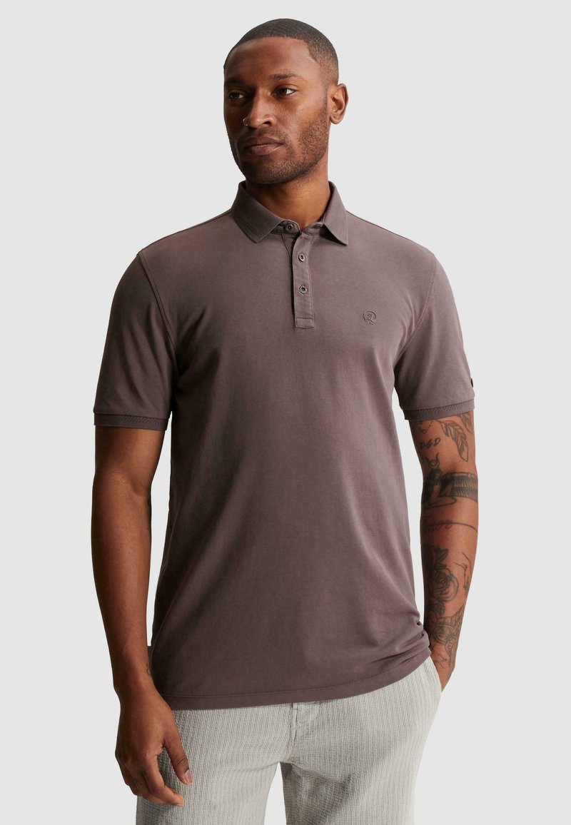 Homme avec des tatouages portant un polo marron à manches courtes et un pantalon clair, debout avec une main dans la poche, regardant vers la gauche.