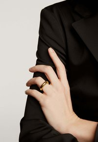 Anillo de metal dorado con un diseño suave y redondeado, llevado en una mano que descansa sobre una superficie de tela negra. Textura sencilla, estilo minimalista.