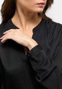 Schwarze, leichte Bluse mit V-Ausschnitt und langen Ärmeln. Glatte Textur mit einem Knopfdetail am Ärmelabschluss, gestaltet für eine taillierte Passform.