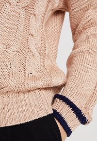 Maglione lavorato a maglia di un rosa chiaro con un motivo a cavo testurizzato, strisce blu navy in contrasto ai polsini. Fondo e polsini a coste, filato morbido.