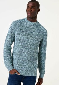 Man in een gestructureerde blauw-groene gebreide trui en donkere spijkerbroek, hand in de zak, naar links kijkend tegen een effen achtergrond.