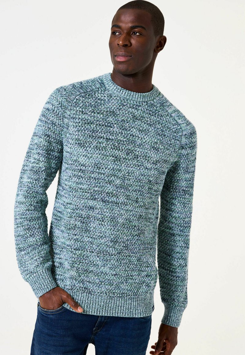 Man in een gestructureerde blauw-groene gebreide trui en donkere spijkerbroek, hand in de zak, naar links kijkend tegen een effen achtergrond.