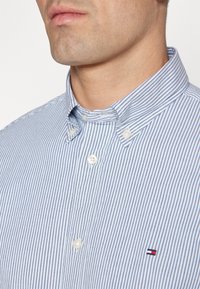 Camicia a righe blu e bianche con colletto e design a bottoni, dotata di una piccola patch con logo sul petto e bottoni perlati.