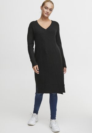 Svart stickad tunika klänning med V-ringning, långa ärmar och sidoflikar, tillverkad av mjukt material, kombinerad med blå jeans och vita sneakers.