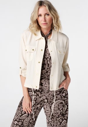 Femme blonde portant une veste crème boutonnée sur une combinaison à motifs floraux marron et blanc, debout les mains dans les poches.