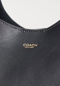 Produit en cuir noir avec une texture lisse, des bords cousus et un logo doré "COACH NEW YORK" affiché de manière proéminente.