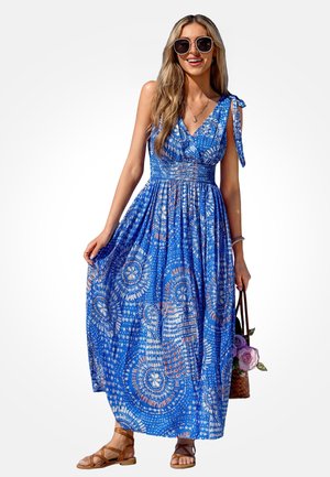 Donna che indossa un vestito lungo senza maniche con motivo blu, sandali marroni, grandi occhiali da sole, che tiene una borsa intrecciata con fiori viola.