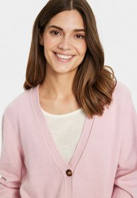 Šviesiai rožinė megztinė su V formos iškirpte, turintis vieną rudą mygtuką ir minkštą tekstūrą, uždėtas ant šviesaus atspalvio marškinėlių.
