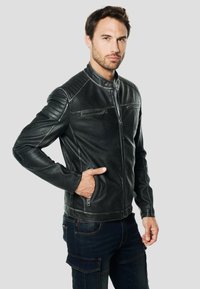 Zwarte leren jas met een opstaande kraag, ritssluiting aan de voorkant en twee ritssluitingen op de borstzakken; voorzien van geribbelde schouderaccenten en gestructureerde mouwen.